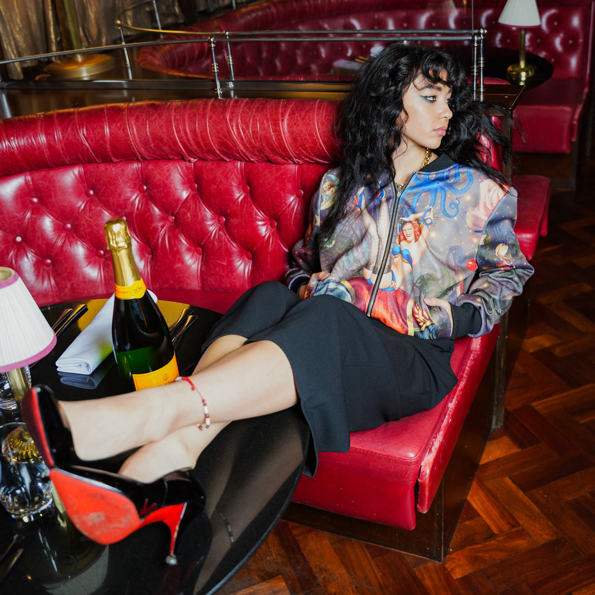 A female model sitting on a sofa wearing luxury softshell bomber jacket in a maximalist retro pinup design 