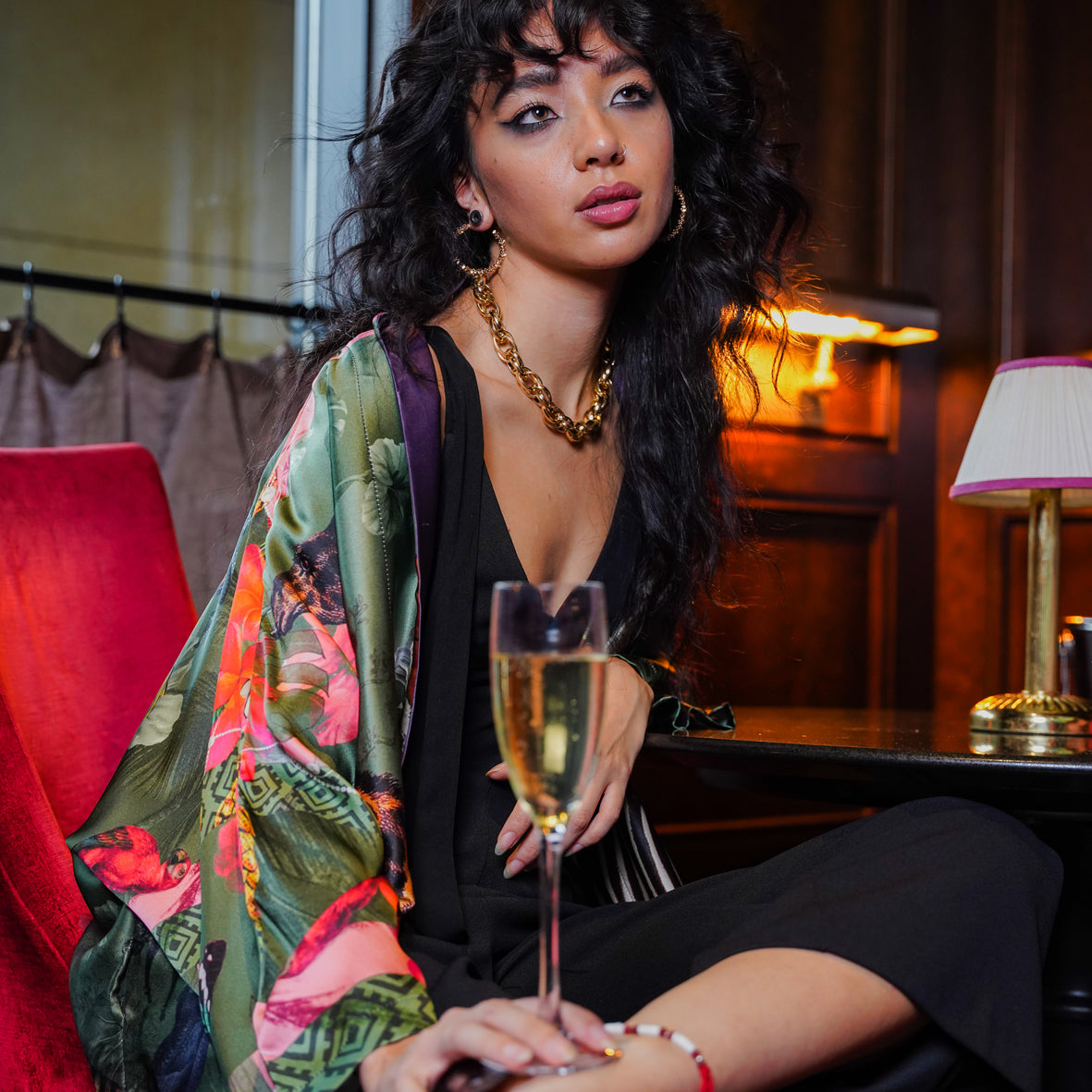 A female model in a bar wearing luxury 100% silk Kimono in a maximalist Carnival inspired design against an olive background 