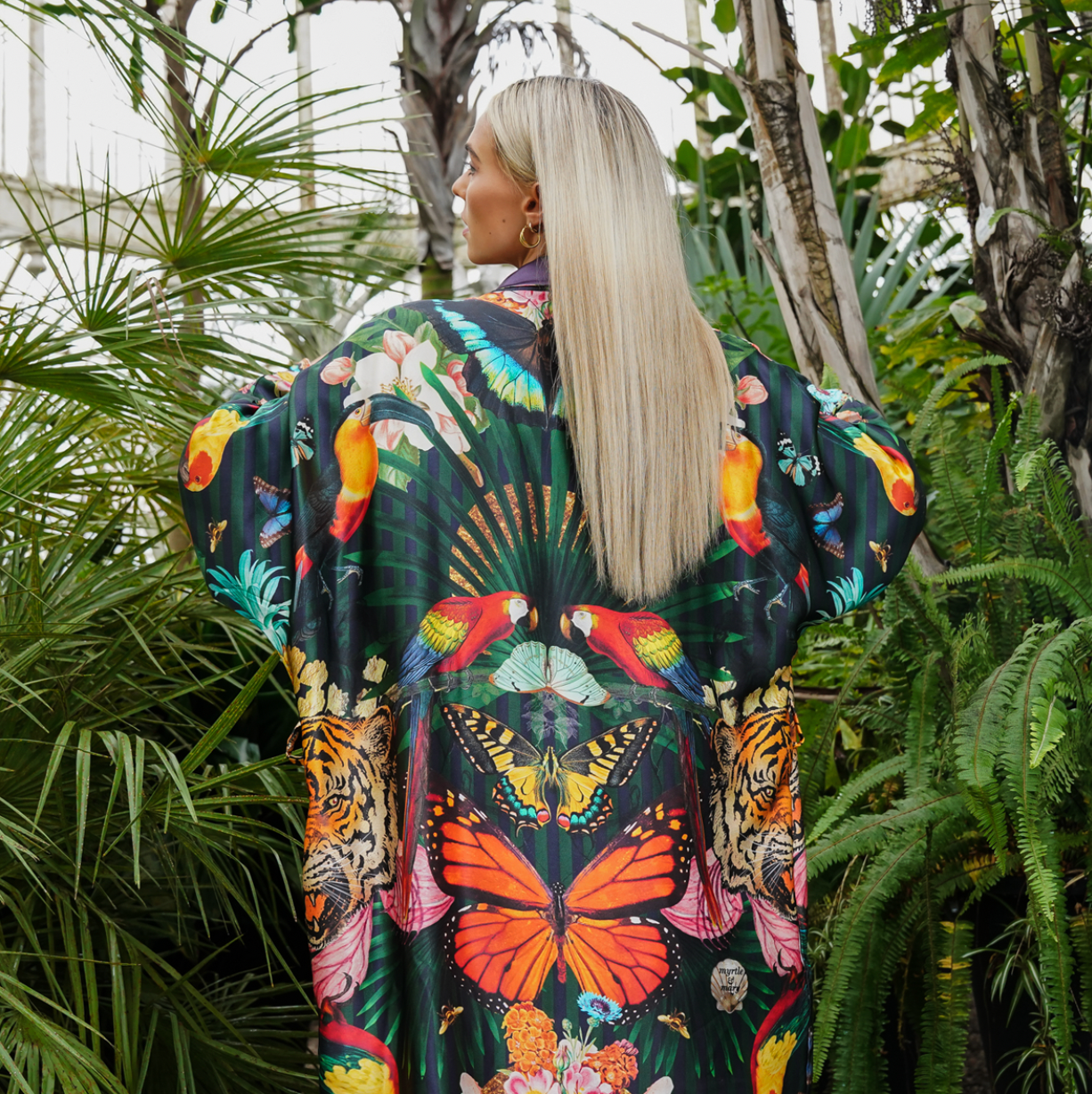 A female model showing the back view of  luxury 100% silk Kimono in a maximalist tropical inspired design against a dark background 