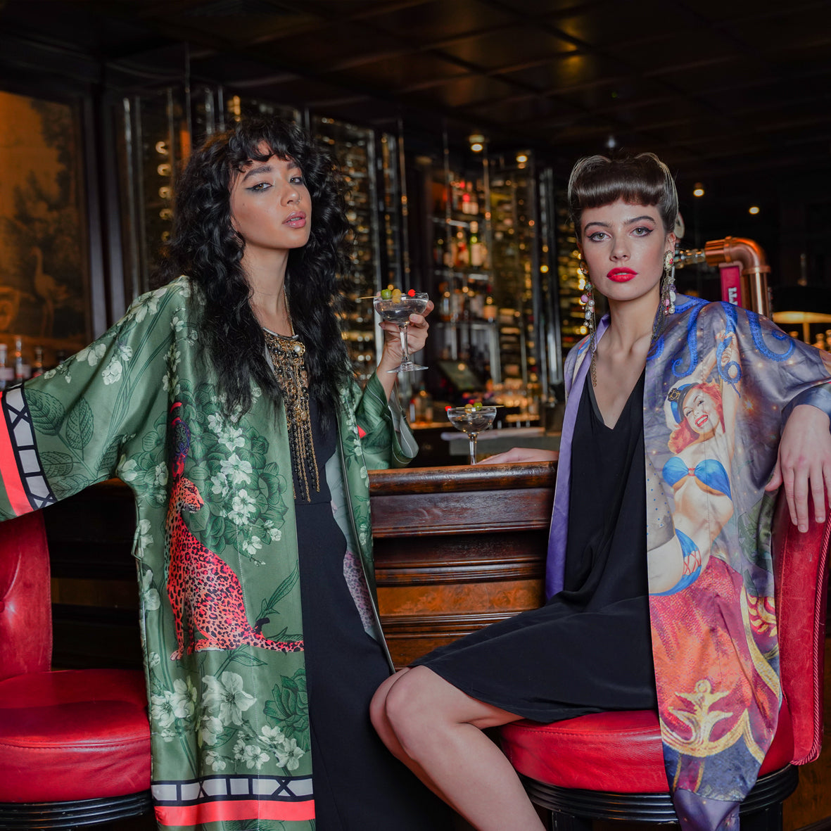 A secondary view of 2 female models sitting in a bar wearing luxury 100% silk kimonos in a maximalist retro pinup design