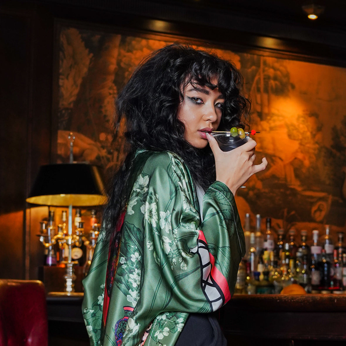 A Female model at a Bar wearing luxury 100% silk Kimono in a green maximalist oriental inspired design 