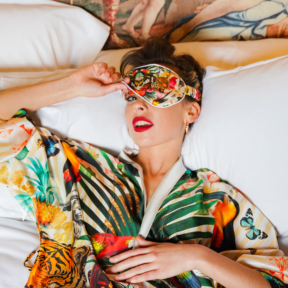 A female model lying on a bed wearing a luxury 100% silk Kimono and matching eye mask in a maximalist tropical inspired design against a pale background 